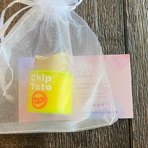 K&ouml;nnte beinhalten: Ein neongelbes "Chip Tato" Seifenst&uuml;ck mit rotem Logo in einem wei&szlig;en Organza-Geschenkbeutel. Die Seife hat eine hellbeige Oberseite, die Kartoffelchips &auml;hnelt. Eine kleine Karte ist hinter dem Beutel sichtbar.