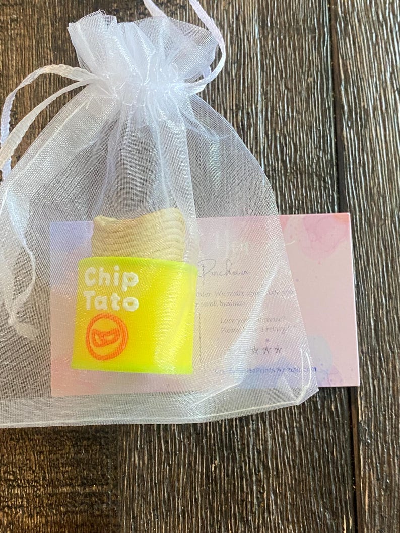 K&ouml;nnte beinhalten: Ein neongelbes "Chip Tato" Seifenst&uuml;ck mit rotem Logo in einem wei&szlig;en Organza-Geschenkbeutel. Die Seife hat eine hellbeige Oberseite, die Kartoffelchips &auml;hnelt. Eine kleine Karte ist hinter dem Beutel sichtbar.