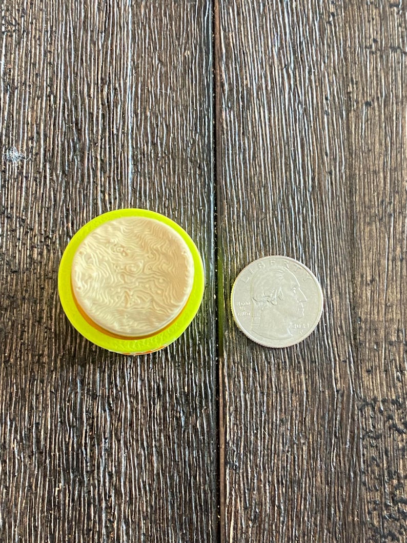 K&ouml;nnte beinhalten: Ein gelb-beiges kreisf&ouml;rmiges Objekt mit strukturiertem Design, neben einer M&uuml;nze. Das Objekt scheint ein Bastel- oder Dekorationsartikel zu sein, m&ouml;glicherweise ein Stempel oder eine Form. Die M&uuml;nze dient als Gr&ouml;&szlig;enreferenz.