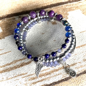 May include: A multi-strand wrap bracelet featuring purple, blue, and light lavender beads interspersed with silver-toned metal spacers. The bracelet includes a silver-toned metal wing charm and a small spiral charm dangling from the strands.