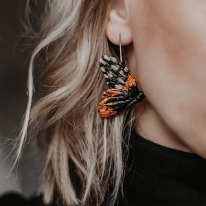 May include: A close-up of a butterfly earring. The earring features a detailed wing design with black, orange, and white patterns. The earring is suspended from a silver hook. The person is wearing a black turtleneck.