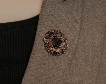 Puede incluir: Blazer marrón con top negro y broche decorativo. El broche es circular con centro oscuro y detalles dorados y marrones. El blazer es de color neutro y tiene un diseño clásico.