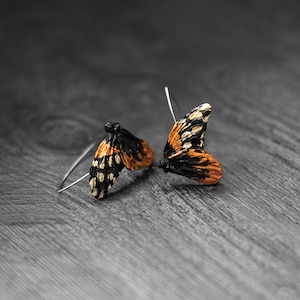 May include: A pair of butterfly wing earrings. The wings are orange, black, and white, with a detailed pattern. The earrings are set on silver hooks, resting on a gray wooden surface.