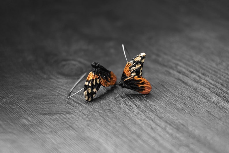 May include: A pair of butterfly wing earrings. The wings are orange, black, and white, with a detailed pattern. The earrings are set on silver hooks, resting on a gray wooden surface.