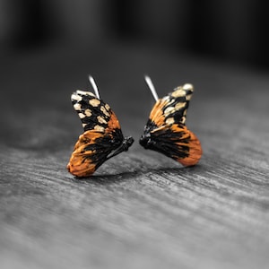 May include: A pair of butterfly wing earrings. The wings are orange, black, and white, with silver earring hooks. The earrings are set against a blurred, grayscale background, highlighting the vibrant colors of the jewelry.