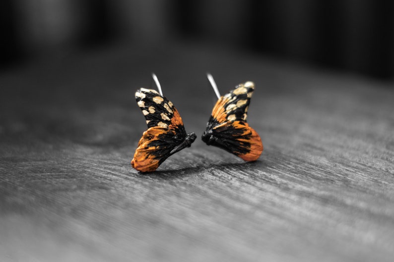 May include: A pair of butterfly wing earrings. The wings are orange, black, and white, with silver earring hooks. The earrings are set against a blurred, grayscale background, highlighting the vibrant colors of the jewelry.