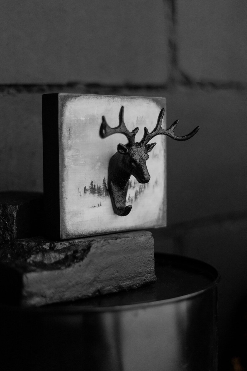 Puede incluir: Gancho de pared de cabeza de ciervo en blanco y negro con un fondo de escena de bosque invernal. El gancho est&aacute; hecho de metal y tiene un aspecto r&uacute;stico.