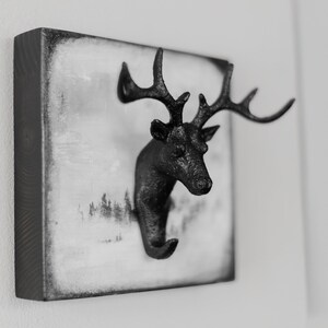 Puede incluir: Gancho de pared de cabeza de ciervo negro con cuernos, montado en una placa de madera cuadrada con fondo blanco y una escena de bosque en blanco y negro.