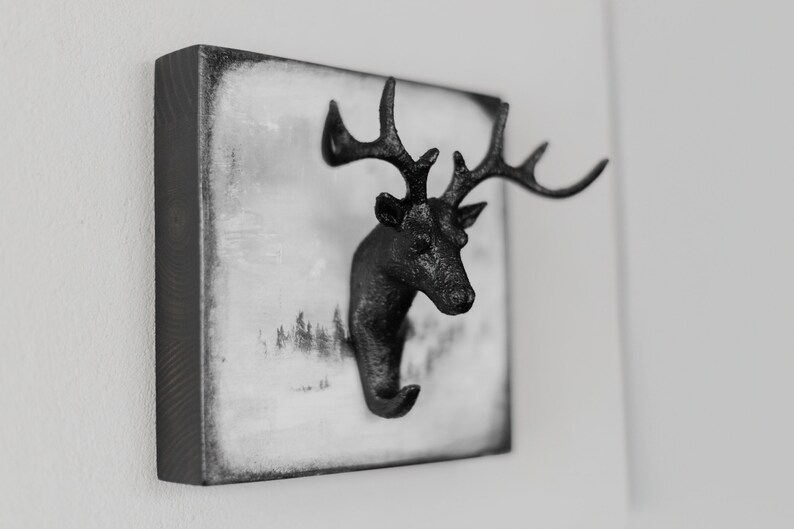 Puede incluir: Gancho de pared de cabeza de ciervo negro con cuernos, montado en una placa de madera cuadrada con fondo blanco y una escena de bosque en blanco y negro.
