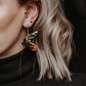 May include: A close-up of a butterfly wing earring. The earring features a detailed design with orange, black, and white colors. The earring is hanging from a silver hook. The background is dark.