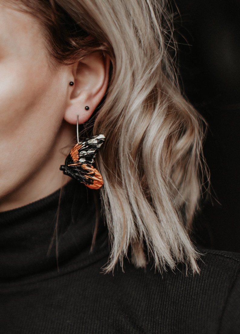 May include: A close-up of a butterfly wing earring. The earring features a detailed design with orange, black, and white colors. The earring is hanging from a silver hook. The background is dark.