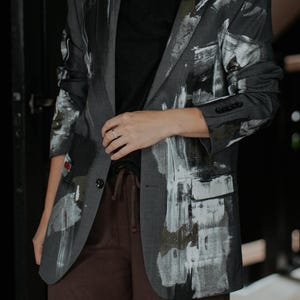 Könnte beinhalten: Ein anthrazitfarbener Blazer mit auffälligen, abstrakten Pinselstrichmustern in Weiß und Dunkelolivgrün auf der Vorderseite und den Ärmeln. Die Jacke hat ein steigendes Revers, einen Knopfverschluss und drei kleine Knöpfe an jedem Ärmel.