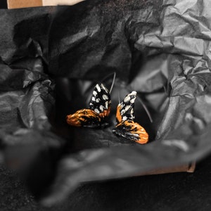 May include: A pair of butterfly-shaped earrings with black and white patterned wings and orange and gold-colored bodies. The earrings are presented on black tissue paper inside a cardboard box, creating a striking contrast.
