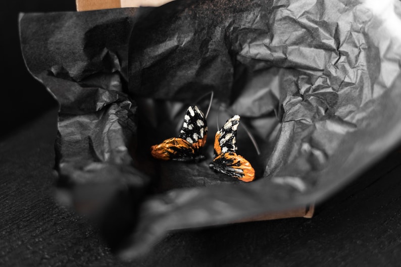 May include: A pair of butterfly-shaped earrings with black and white patterned wings and orange and gold-colored bodies. The earrings are presented on black tissue paper inside a cardboard box, creating a striking contrast.