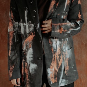 Puede incluir: Una chaqueta tipo blazer negra con patrones abstractos de pinceladas en pintura color cobre y blanca. La prenda incluye solapas, dos botones frontales, dos bolsillos inferiores y pequeñas siluetas de aves blancas pintadas en el frente.