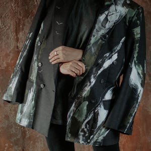 May include: A dark charcoal grey double-breasted blazer featuring an abstract painted design in shades of white, forest green, and black. The blazer has structured shoulders, notched lapels, and dark buttons, with small white bird silhouettes.