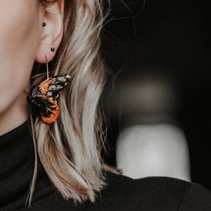 May include: Close-up of a butterfly wing earring. The earring features a black, orange, and white wing design. The earring is attached to a silver hoop. The person is wearing a black turtleneck.