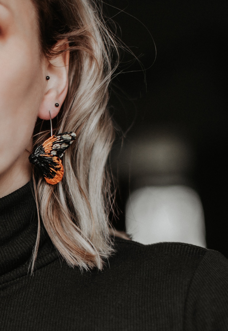 May include: Close-up of a butterfly wing earring. The earring features a black, orange, and white wing design. The earring is attached to a silver hoop. The person is wearing a black turtleneck.