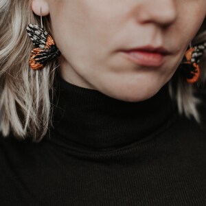 May include: Close-up of a person wearing moth-shaped earrings. The earrings feature intricate black, orange, and white details, with a silver hook. The person is wearing a black turtleneck sweater, and has blonde hair.