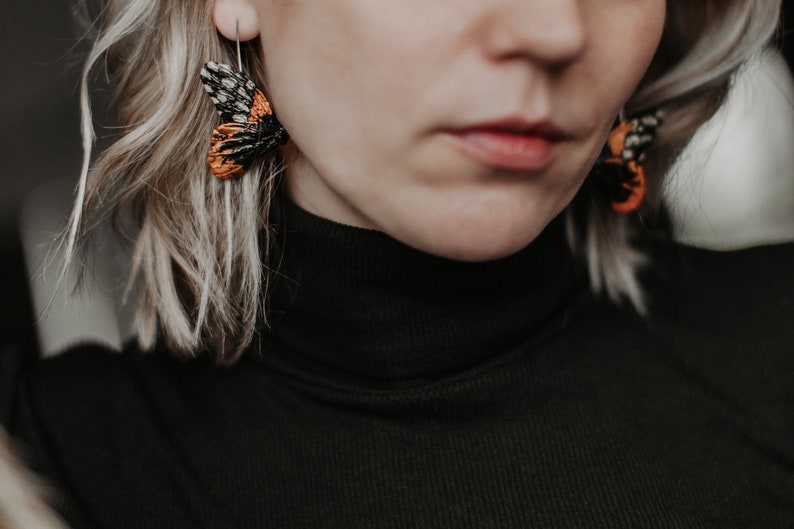 May include: Close-up of a person wearing moth-shaped earrings. The earrings feature intricate black, orange, and white details, with a silver hook. The person is wearing a black turtleneck sweater, and has blonde hair.