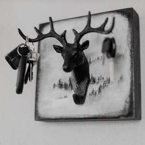 Puede incluir: Porta llaves de cabeza de ciervo negro sobre fondo blanco con una escena de invierno. La cabeza de ciervo tiene cuernos y un gancho para colgar llaves. Hay llaves colgando del gancho.