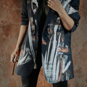 Pode incluir: Um blazer azul-marinho com riscas de giz, apresentando pinceladas arrojadas pintadas à mão em tons de branco, preto e cobre na parte frontal e nas mangas. O blazer tem lapela entalhada, dois bolsos com aba e botões na parte frontal.