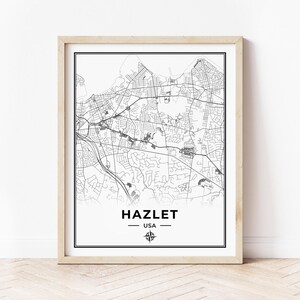 May include: A framed black and white map print of Hazlet, USA. The map shows detailed street layouts and geographical features. The town's name is centered at the bottom, with a decorative line and compass rose symbol. The print is in a light-coloured wooden frame.