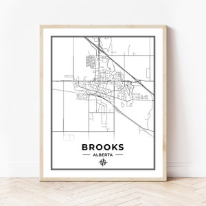 May include: A framed black and white map print of Brooks, Alberta. The map features a detailed street layout with the city name and province at the bottom. The print is in a light-colored wooden frame.