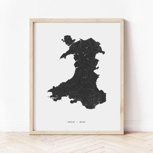 May include: A framed print featuring a detailed black and white map of Wales. The map is centered on a white background, with the country's outline and internal features rendered in black. The frame is a light wood color, and the text "WEALAS | WALES" is at the bottom.