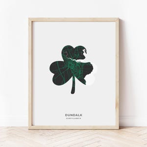 Może przedstawiać: Mapa Dundalk w Irlandii w kształcie koniczyny w kolorze czarnym i zielonym, z nazwą miasta i współrzędnymi nadrukowanymi poniżej. Mapa jest oprawiona w jasną drewnianą ramę i wyświetlana na białym tle.