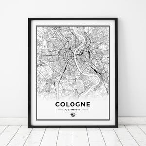 Puede incluir: Impresión de mapa en blanco y negro de Colonia, Alemania. El mapa muestra las calles de la ciudad y el río Rin. La impresión tiene un marco negro y un símbolo de brújula en el centro inferior.