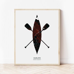May include: A black and red map of Guelph, Ontario, Canada, is printed on a canoe paddle with two white paddles crossed behind it. The text "GUELPH" and "43.5448° N, 80.2482° W" is printed below the canoe paddle.