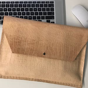 May include: A light brown leather laptop sleeve with a crocodile-embossed pattern. The sleeve has a flap closure with a single snap button.