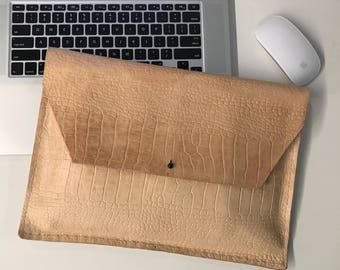 May include: A light brown leather laptop sleeve with a crocodile-embossed pattern. The sleeve has a flap closure with a single snap button.
