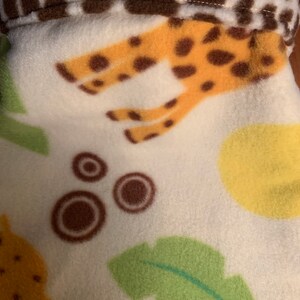 May include: White fleece fabric with a brown and yellow giraffe print, green leaves, and brown circles.