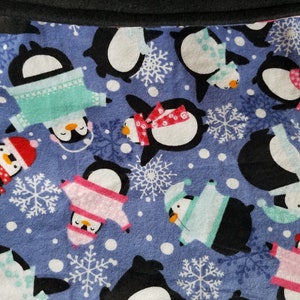 May include: Close-up of a blue fabric with a pattern of penguins wearing winter clothing and snowflakes. The penguins are black and white with orange beaks, and the clothing is pink, blue, and red. The top edge is a dark gray.