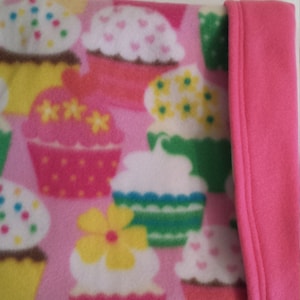 May include: Pink fleece blanket with a cupcake pattern. The blanket has a pink fleece border.