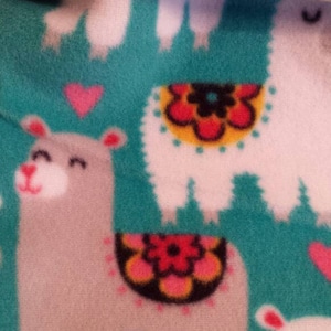 May include: A teal blue fleece fabric with a white background featuring a repeating pattern of llamas with colorful flowers and hearts. The llamas are gray and white with black eyes and pink noses.