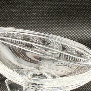 May include: Clear glass serving dish with a decorative, oval-shaped bowl. The dish has a pedestal base and intricate etched patterns along the rim. The glass is transparent, allowing light to pass through.