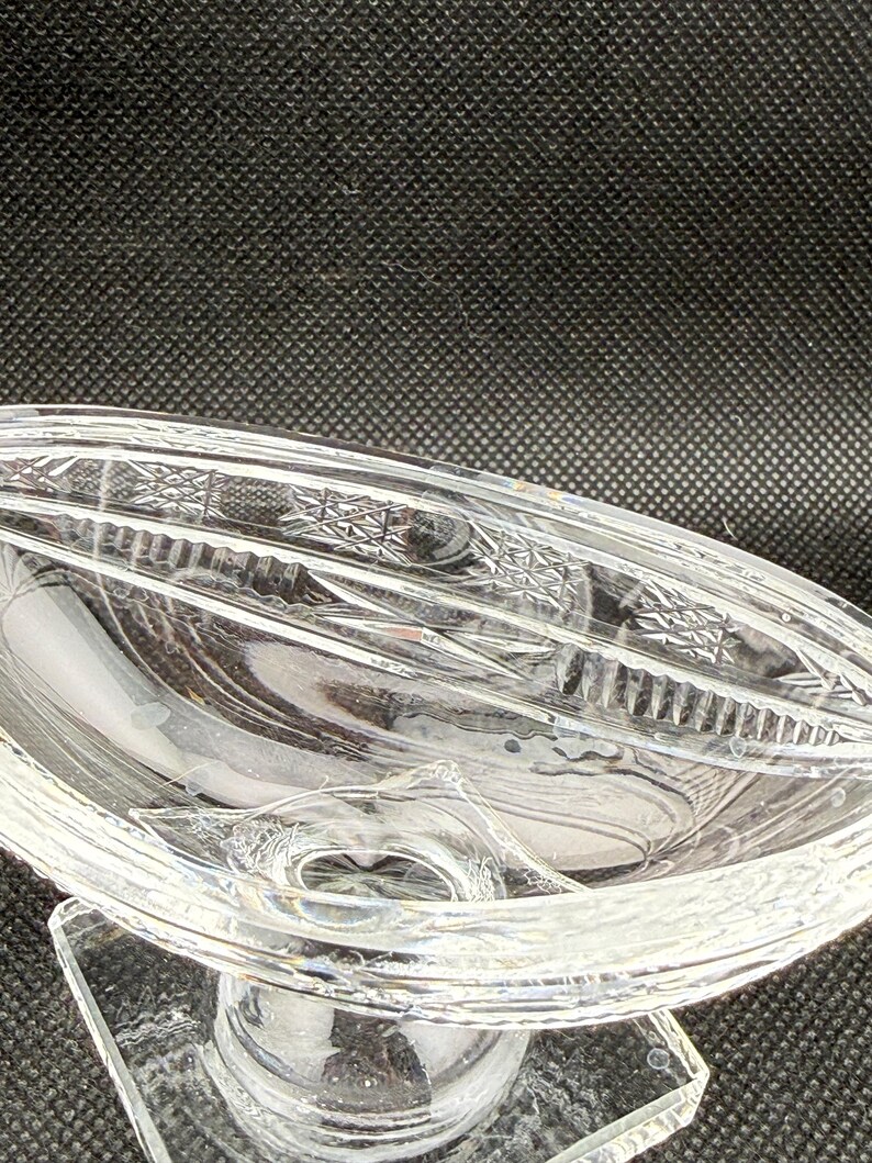 May include: Clear glass serving dish with a decorative, oval-shaped bowl. The dish has a pedestal base and intricate etched patterns along the rim. The glass is transparent, allowing light to pass through.