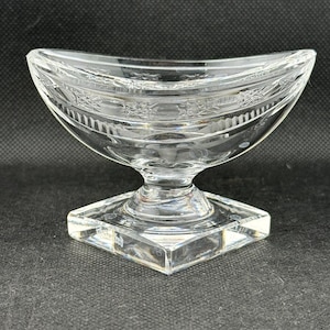 May include: Clear glass serving dish with a curved bowl and a square base. The dish has etched floral patterns around the rim and a pedestal base. The glass is transparent, allowing light to pass through.