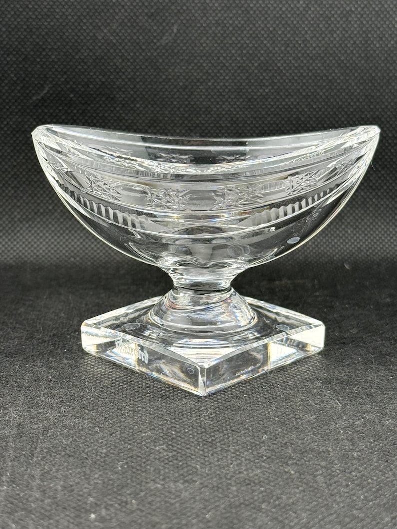 May include: Clear glass serving dish with a curved bowl and a square base. The dish has etched floral patterns around the rim and a pedestal base. The glass is transparent, allowing light to pass through.