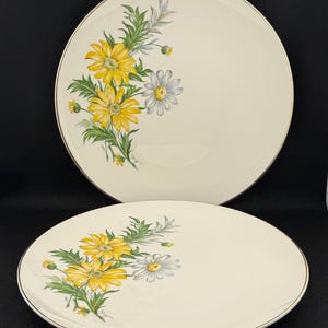 May include: Two cream-coloured ceramic plates with a floral design. The plates feature yellow and white daisies with green leaves and a gold rim. The plates are round and are likely for serving food.