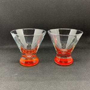 May include: Two clear martini glasses with vibrant orange bases. The glasses have a classic conical bowl shape and a short, rounded stem. The transparent design allows the color of the drink to be visible. Perfect for cocktails and other beverages.