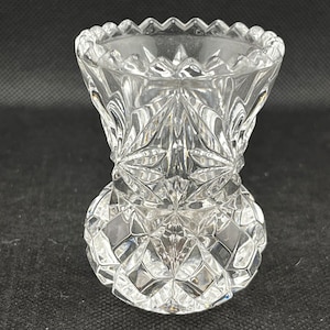 May include: A clear glass bud vase with a cut glass design. The vase has a scalloped rim and a diamond-shaped pattern.