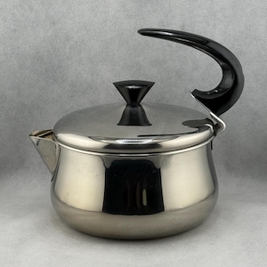 May include: A stainless steel teapot with a black handle and lid knob. The teapot has a curved handle and a spout for pouring. The lid sits on top of the pot. The teapot is reflective and appears to be in good condition.