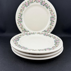 May include: A stack of three white ceramic plates with a floral border, and one plate on top. The floral design includes pink, blue, and yellow flowers with green leaves. The plates are on a black surface.