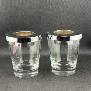 May include: Two clear glass votive candle holders with a silver-colored rim and a gold-colored interior. The candle holders are cylindrical and sit on a dark background.