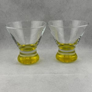 May include: Two clear glass martini glasses with yellow bases. The glasses are empty and sitting on a white surface.