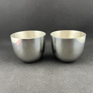 May include: Two small, silver-colored metal cups with a rounded, bowl-like shape. The cups have a smooth, shiny interior and a slightly textured exterior. The cups are displayed on a gray surface.